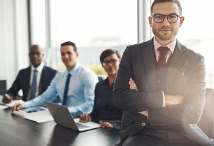 bearded professional man sitting on desk corner beside three coworkers in front of large office window | association management